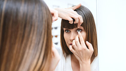 Frau vor einem Spiegel, die sich gerade eine Kontaktlinse einsetzt