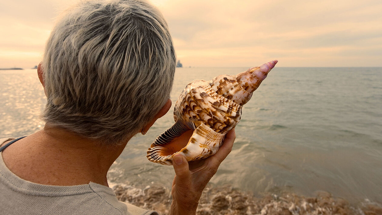 Eine ältere Dame lauscht am Meer an einer Muschel